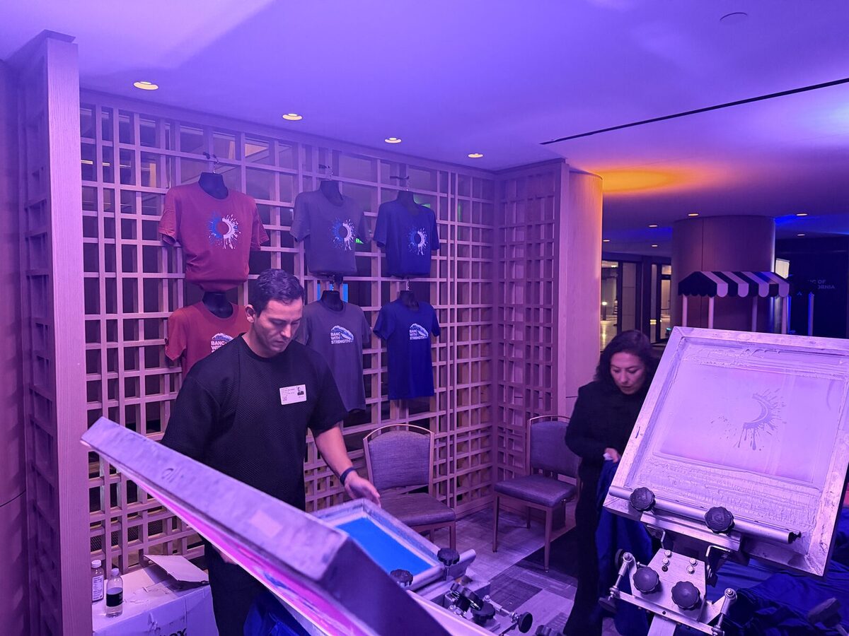 Two Merch Troop printers loading screens on a press with freshly printed tees hanging behind them