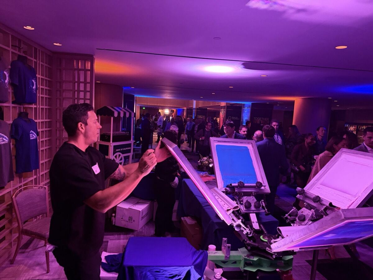 Two Merch Troop printers loading screens on a press with freshly printed tees hanging behind them