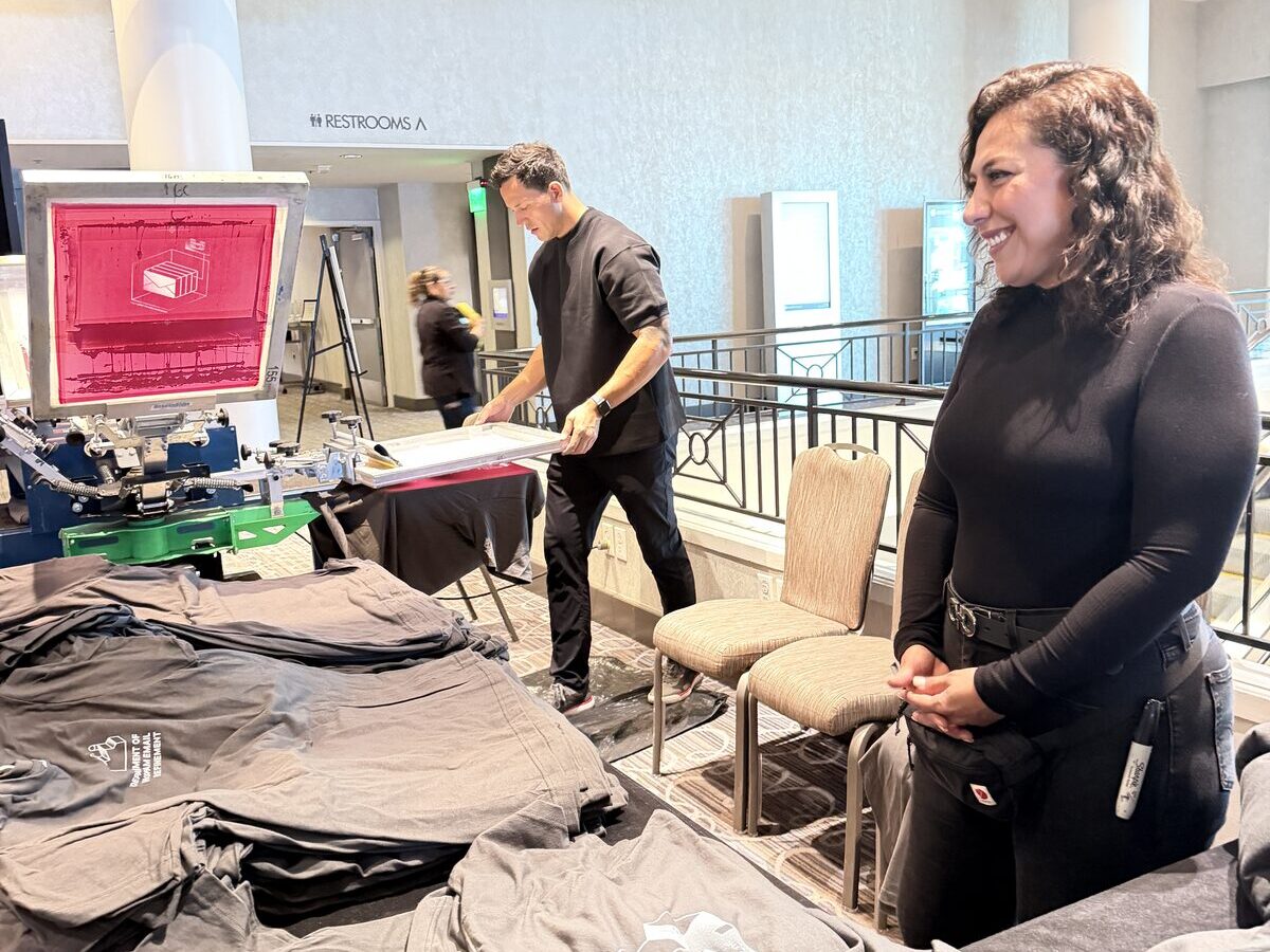 Two smiling Merch Troop staff at a bright printing station beside a loaded press and stacks of folded printed tees